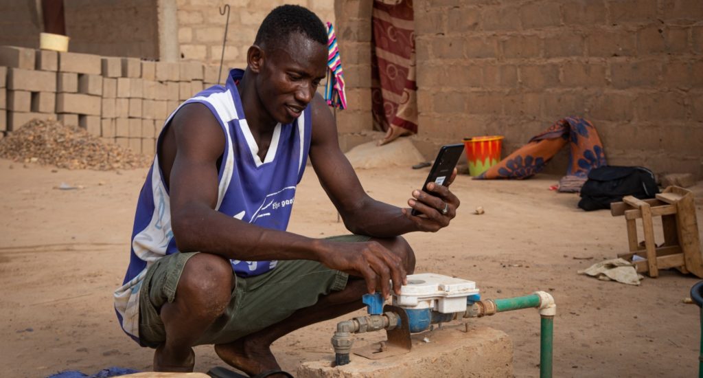 A man kneeling, looking at a mobile phone and a water pipe and tap