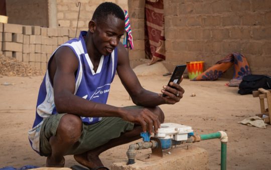 A man kneeling, looking at a mobile phone and a water pipe and tap
