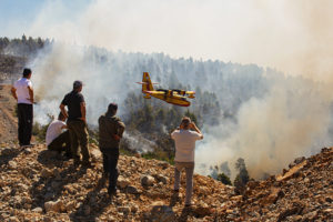 Locals look at burning forest during a wildfire. A plane is flying low, possibly preparing to drop water