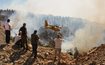 Locals look at burning forest during a wildfire. A plane is flying low, possibly preparing to drop water