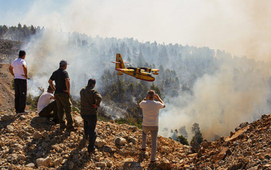 Locals look at burning forest during a wildfire. A plane is flying low, possibly preparing to drop water