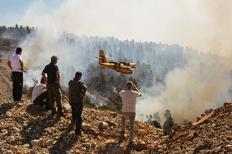 Locals look at burning forest during a wildfire. A plane is flying low, possibly preparing to drop water