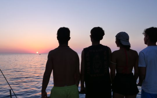 Four figures in silhouette, looking out over the sea at sunset