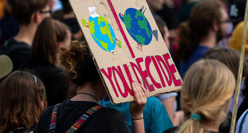 Someone in a crowd holding up a sign that says "you decide" with the image of two versions of Planet Earth depicted - a brighter one and a darker one