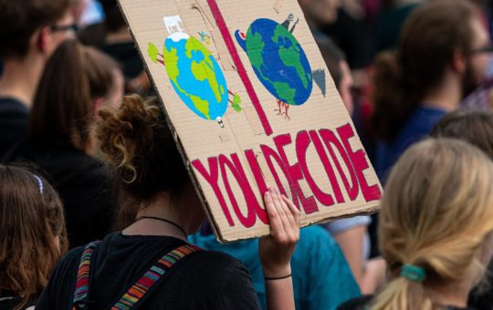 Someone in a crowd holding up a sign that says "you decide" with the image of two versions of Planet Earth depicted - a brighter one and a darker one