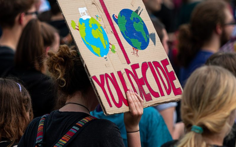 Someone in a crowd holding up a sign that says "you decide" with the image of two versions of Planet Earth depicted - a brighter one and a darker one