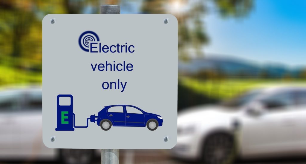 A sign that says "electric vehicles only" with an image of a car and a charger. Two cars are visible in the background