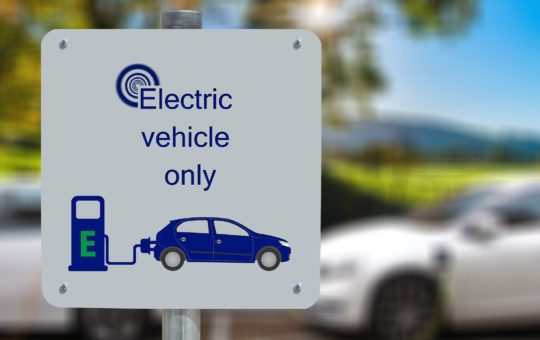 A sign that says "electric vehicles only" with an image of a car and a charger. Two cars are visible in the background