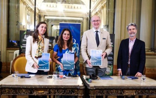 Gabriella Seiler, Malini Mehra, Stuart Brocklehurst, Carlos Milani standing behind a table in a large a grand room, holding copies of the climate guide