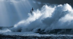 A large wave breaking on a rocky coast, with a lighthouse just visible in the background