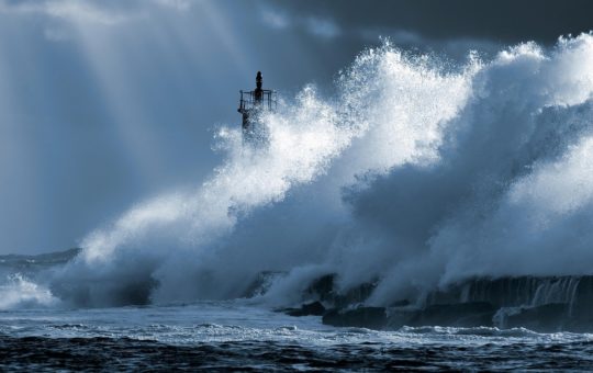 A large wave breaking on a rocky coast, with a lighthouse just visible in the background