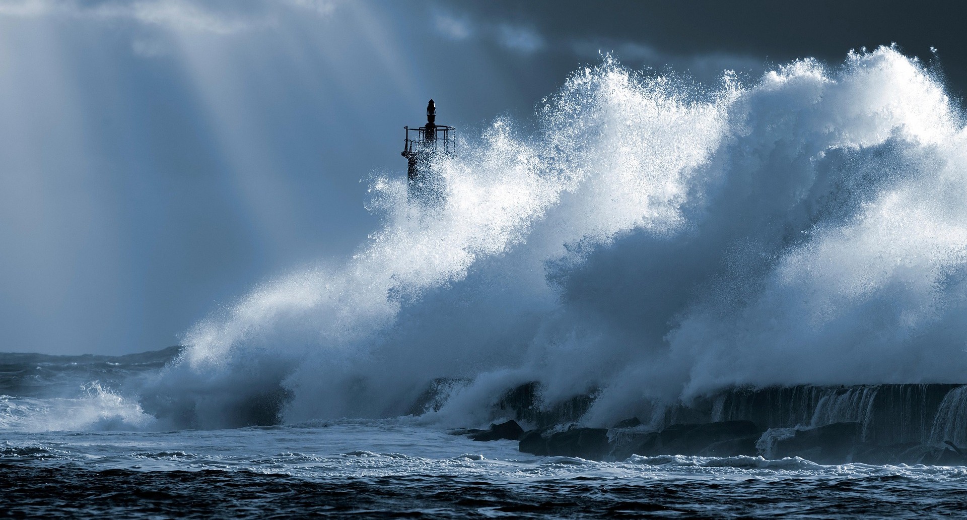 A large wave breaking on a rocky coast, with a lighthouse just visible in the background