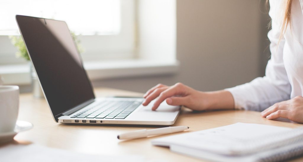 A person sitting at a laptop, with their hand on the keyboard. A notebook is visible on the table