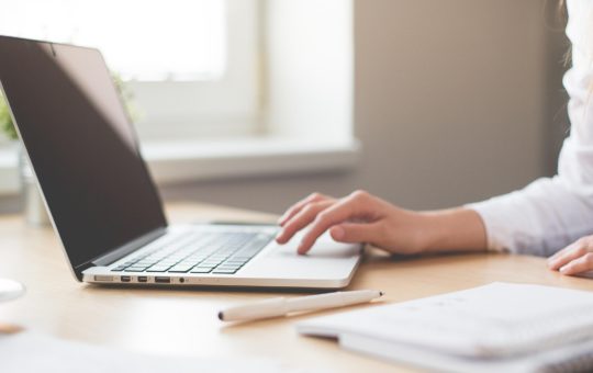 A person sitting at a laptop, with their hand on the keyboard. A notebook is visible on the table