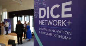 A closeup of a sign that says DICE Network+ with some blurred people visible in a meeting room in the background
