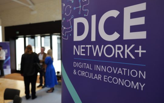 A closeup of a sign that says DICE Network+ with some blurred people visible in a meeting room in the background