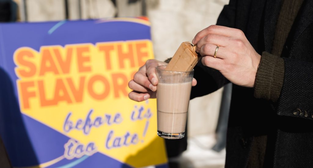 A close-up of someone's hands, dipping a biscuit into a chocolate drink in a glass. A sign in the background says "save the flavors"