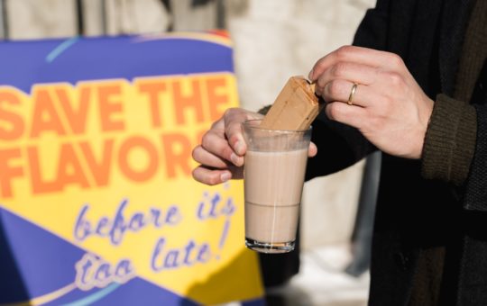 A close-up of someone's hands, dipping a biscuit into a chocolate drink in a glass. A sign in the background says "save the flavors"