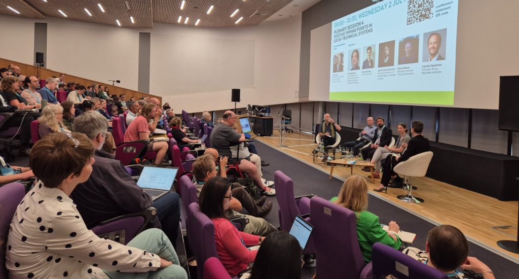 A speaker and a panel on stage in the Alumni Auditorium at the University of Exeter, with an audience watching