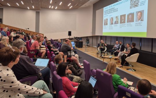 A speaker and a panel on stage in the Alumni Auditorium at the University of Exeter, with an audience watching