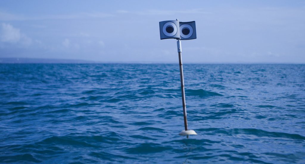 A pole mounted on a buoy at sea, with a spinning metal top displaying a pair of large eyes on each side