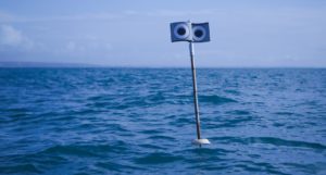 A pole mounted on a buoy at sea, with a spinning metal top displaying a pair of large eyes on each side